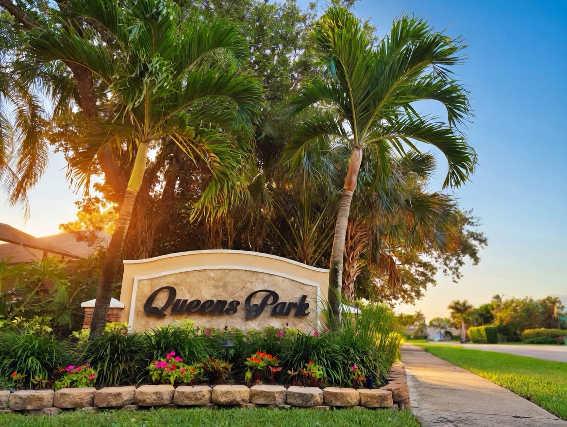 The Queen's Park entrance monument at golden hour — a low stone sign lettered 'Queens Park', framed by royal palms and tropical plantings in East Naples.