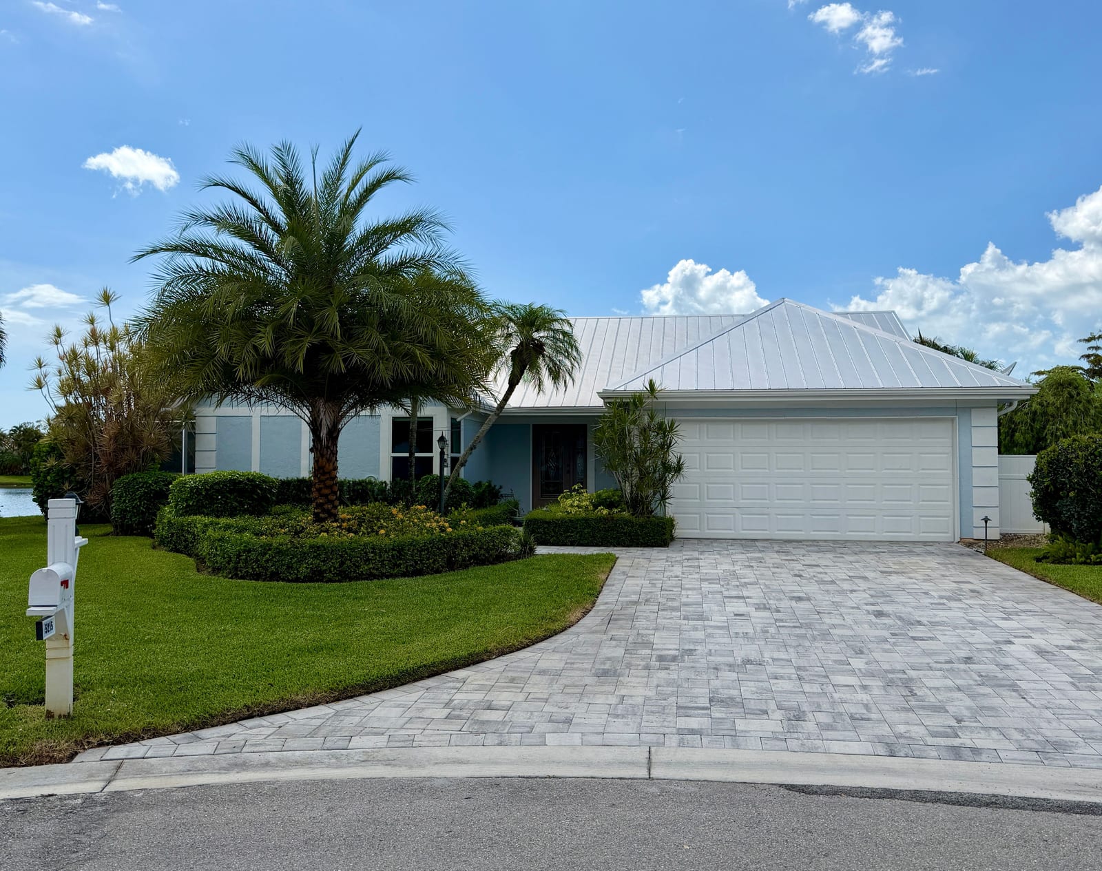 A single-story Queen's Park home on a lakefront lot in East Naples — pale-blue exterior, white standing-seam metal roof, mature palms, and a paver driveway.