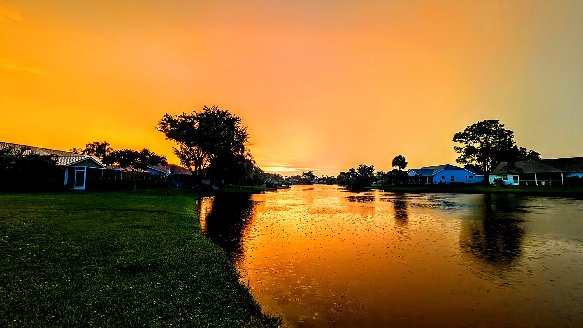 Queen's Park interior lake at sunset — silhouetted oaks and palms framing single-family lanais along the shoreline, with an orange Florida sky reflecting on rain-rippled water after a summer storm.